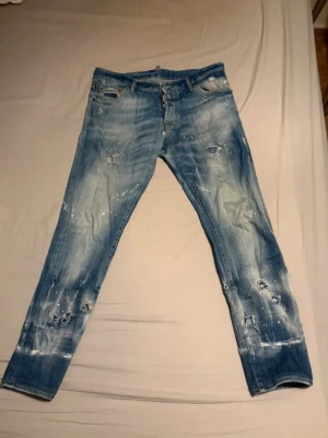 Dsquared 2 storlek 50/S/M - Säljer ett par blå jeans från Dsquared2 med slitningar, hål och vita färgstänk för en riktigt trendig och avslappnad look. Jeansen har klassisk femficksmodell och knappgylf. Perfekta för dig som gillar streetwear och vill sticka ut.