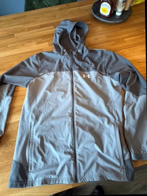 Grå vindjacka från Under Armour - Snygg grå vindjacka från Under Armour med huva och dragkedja framtill. Jackan har logga på bröstet och tryckt text på ärmslutet. Tillverkad i lätt och slitstarkt material, perfekt för blåsiga dagar.aldrig använd 