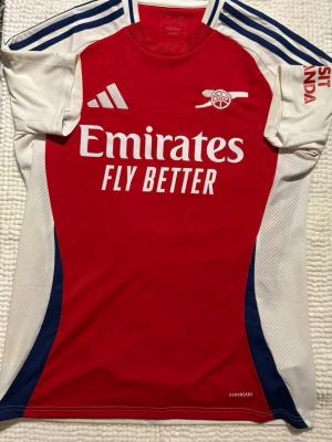Arsenal matchtröja röd/vit Adidas 2025/2026 - Säljer en Arsenal matchtröja från Adidas i rött och vitt med blå detaljer. Tröjan har klubbmärke och Adidas-logga på bröstet samt 'Emirates Fly Better' tryckt framtill. Tillverkad i lätt och ventilerande material med AEROREADY-teknologi.