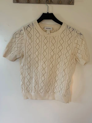 Vit stickad t-shirt från Monki - Vit stickad t-shirt från Monki med korta ärmar och ett snyggt hålmönster i diamantform. Tröjan har rund halsringning och ribbade muddar vid ärmslut och nederkant. Perfekt för lager-på-lager och en clean look.