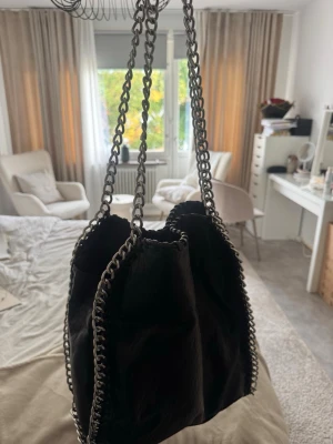 Svart kedjehandväska från tiamo - Cool svart handväska från tiamo med silverfärgade kedjedetaljer längs kanterna och som axelrem. Väskan är tillverkad i konstläder och har ett rymligt huvudfack med dragkedja samt innerficka. Perfekt för dig som gillar en edgy och trendig look.