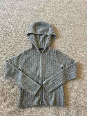 Grå ribbstickad hoodie med dragkedja - Mysig grå hoodie i ribbstickat material med dragkedja framtill och huva. Tröjan har långa ärmar och en avslappnad passform, perfekt för chill dagar. Materialet ser ut att vara mjukt och stretchigt, vilket gör den extra bekväm.