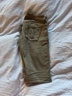 Olivgröna bootcut jeans med brodyr - Säljer ett par olivgröna bootcut jeans med snygg broderad ficka bak och slitning på ena låret. Jeansen har två knappar fram och vida ben. Perfekta för dig som gillar en avslappnad men trendig stil.