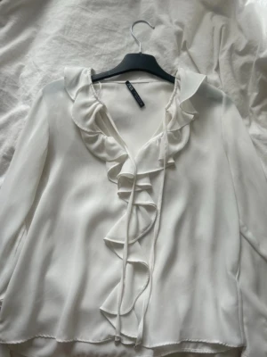 Vit volangblus med långa ärmar - Säljer en elegant vit blus från Zara med långa ärmar och volangdetaljer längs ringningen och framtill. Blusen är tillverkad i ett tunt, lätt material som ger ett luftigt intryck. Perfekt för dig som gillar en romantisk och stilren look.