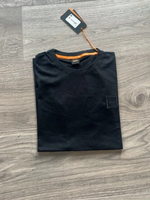 Svart t-shirt från BOSS - Snygg svart t-shirt från BOSS med diskret logga på bröstet. Klassisk rund halsringning och korta ärmar. Tillverkad i mjuk bomull som känns skön mot huden. Perfekt basplagg med stilrena detaljer och orange insida vid halsen.
