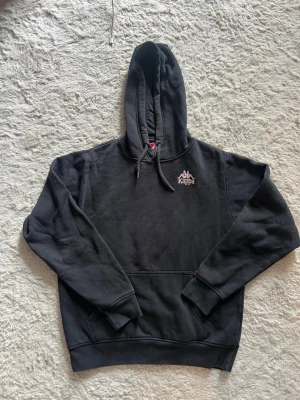 Svart hoodie från Kappa med rosa tryck - Säljer en svart hoodie från Kappa med stor rosa logga på ryggen och mindre logga på bröstet. Klassisk känguruficka framtill, ribbade muddar och justerbar huva med snören. Perfekt för en chill och sportig look.