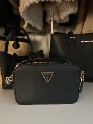 Svart crossbody-väska från Guess - Snygg svart crossbody-väska från Guess i slätt konstläder med guldfärgade detaljer och kedjerem. Väskan har två dragkedjefack och den klassiska Guess-loggan framtill. Perfekt storlek för det viktigaste och ger en trendig touch till din outfit. Priset kan diskuteras!