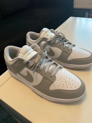 Nike Dunk Low gråvita sneakers - Helt nya Nike Dunk Low sneakers i vitt och ljusgrått läder. Klassisk siluett med perforerad tåbox, vadderad plös och matchande grå skosnören. Perfekta för dig som gillar stilrena och tidlösa sneakers med en sportig vibe. Hör av dig vid frågor!