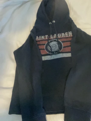 Mörkblå hoodie med tryck - Snygg mörkblå hoodie med huva och dragsko. På bröstet finns ett tryck med texten 'SAINT LAURENT' och ett emblem i vitt, rött och blått. Hoodien har långa ärmar och ribbade muddar. Perfekt för en avslappnad och trendig stil.