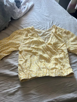 Gul blommig omlottblus - Söt gul omlottblus med små vita blommor och v-ringning. Blusen har långa ärmar med lätt puff och resår i midjan för en snygg passform. Perfekt för dig som vill ha en fräsch och somrig look.