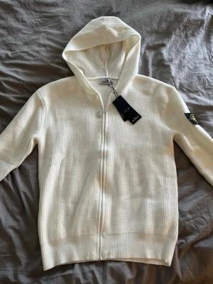 Vit hoodie med dragkedja från Stone Island - Säljer en stilren vit hoodie från Stone Island med dragkedja framtill och ribbad struktur. Hoodien har en klassisk huva och den ikoniska Stone Island-loggan på vänster ärm. Perfekt för dig som gillar exklusiva streetwear-plagg. S/M