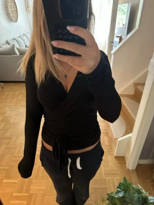 Svart omlottopp med knyt - Snygg svart omlottopp med långa ärmar och knyt i midjan. Toppen har en v-ringad design och sitter tight för en smickrande siluett. Perfekt att styla med både jeans och kjol för en trendig look.