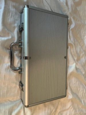 Poker set i aluminiumväska - Snyggt pokerset i en silverfärgad aluminiumväska med räfflad yta. Innehåller marker i grönt, svart, blått, rött och vitt, en dealerknapp samt en kortlek. Perfekt för pokerkvällar med vänner. Väskan har metallhandtag och lås för enkel transport.