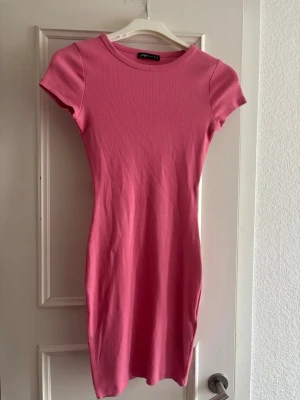 Rosa ribbad kortklänning - En enkel och snygg rosa kortklänning med ribbad struktur och rund hals. Klänningen har korta ärmar och en kroppsnära passform som framhäver siluetten. Perfekt för varma dagar och kan stylas på många olika sätt.