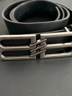 Svart bälte med unik metallspänne - Snyggt svart bälte i skinn med ett coolt, geometriskt metallspänne i silverfärg. Perfekt accessoar för att lyfta din outfit med en modern touch. Passar till jeans eller byxor och ger en stilren look.