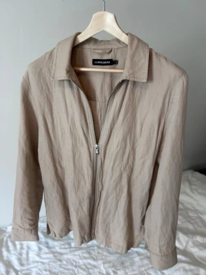 Beige overshirt från J.Lindeberg - Stilren beige overshirt från J.Lindeberg med dragkedja framtill och klassisk krage. Tillverkad i ett lätt, något skrynkligt material som ger en avslappnad look. Perfekt att bära över en t-shirt för en enkel men snygg outfit.