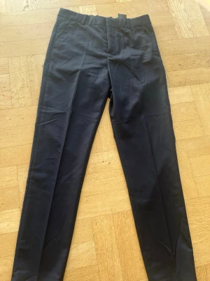 Svarta klassiska kostymbyxor - Snygga svarta kostymbyxor med raka ben och pressveck. Byxorna har hällor för bälte och en bakficka med knapp. Perfekta för en stilren look och passar till skjorta eller t-shirt.                            Storlek 170