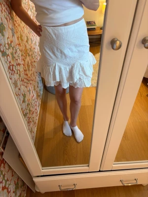Vit kortkjol med volang och spets -  Vit kortkjol med bred volangkant och fina spetsdetaljer. Kjolen har en feminin och somrig look med figurnära passform upptill och lite utsvängd nertill. 