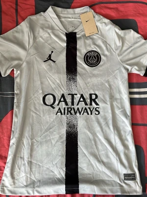 PSG vit matchtröja Nike Dri-FIT - Säljer en vit Paris Saint-Germain matchtröja från Nike med svart vertikal rand och Jordan-logga. Tröjan har PSG-emblem på bröstet och Qatar Airways tryckt på framsidan. Tillverkad i lätt och ventilerande Dri-FIT-material, perfekt för fotboll eller träning.