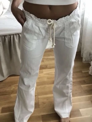 Vita linnebyxor från Roxy - Säljer ett par vita linnebyxor med avslappnad passform och bred resår i midjan. Byxorna har snörning med repdetalj och stora fickor framtill. Perfekta för en chill och stilren look.