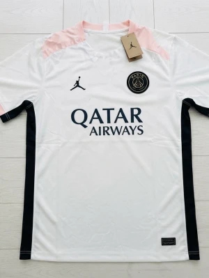 PSG matchställ vit/svart/rosa - Säljer ett PSG matchställ med vit tröja och svarta shorts. Tröjan har rosa axelpartier, svarta detaljer och PSG-logga samt Jordan-logga på bröstet. Shortsen är svarta med rosa inslag och PSG-märke. Materialet är lätt och andas, perfekt för fotbollsträning.