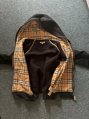 Svart hoodie med klassiskt rutigt foder - Snygg svart hoodie med dragkedja och huva. Insidan har det ikoniska beige, röda och svarta rutmönstret. Utsidan är helsvart och materialet ser ut att vara mjukt och bekvämt, perfekt för en chill look. Dragkedjan är i metall och känns rejäl. Storlek S men passar M.Jag säljer endast denna för att den är för liten!