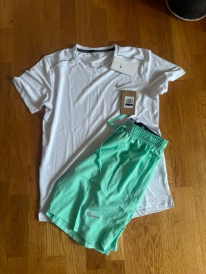 Nike vit t-shirt och mintgröna shorts - Säljer ett sportigt set från Nike med en vit t-shirt i lätt och snabbtorkande material samt ett par mintgröna shorts med resår i midjan och dragsko. Båda plaggen har Nike-logga tryckt på framsidan. Perfekt för träning eller löpning.