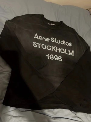 Svart långärmad t-shirt Acne Studios - Cool svart långärmad t-shirt från Acne Studios med vit text 'Acne Studios STOCKHOLM 1996' på bröstet. Klassisk rund halsringning och relaxed fit. Perfekt för dig som gillar stilrena och trendiga plagg.