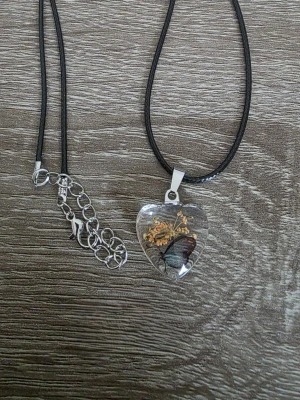 Halsband med hjärthänge och blomma - Unikt halsband med svart vaxad rem och ett hjärtformat hänge i glas. Hänget innehåller en liten torkad blomma och en fjäril, vilket ger en cool och naturinspirerad look. Justerbar kedja i silverfärg för perfekt passform.