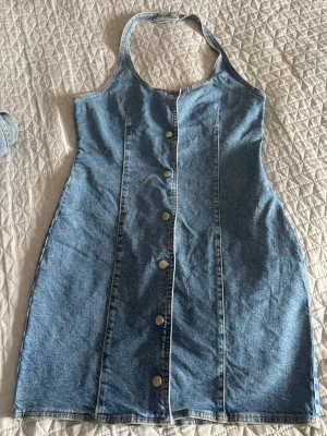 Kort jeansklänning med knappar - Säljer en snygg kort jeansklänning i blå denim med halterneck och knappar framtill. Klänningen har figurnära passform och är ärmlös, perfekt för sommarens alla tillfällen. Klassisk och enkel design som är lätt att styla.