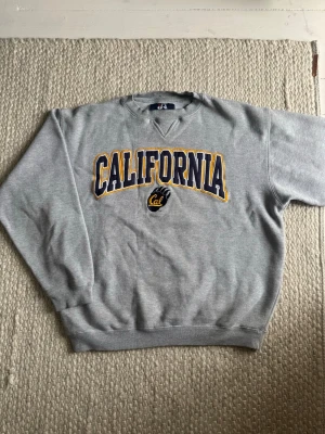 Grå sweatshirt med California-tryck - Snygg grå sweatshirt med stort 'CALIFORNIA' tryck i blått och gult på bröstet samt Cal-logga. Klassisk rund halsringning och ribbade muddar vid ärmslut och nederkant. Perfekt för en avslappnad och sportig stil.