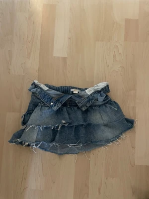 Blå jeanskjol med volanger - Snygg blå jeanskjol med volanger och råa kanter. Kjolen har en kort modell och är tillverkad i denim med slitna detaljer för en trendig look. Perfekt för dig som gillar en avslappnad och cool stil.