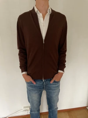 Brun ziptröja i klassisk stil - Mörkbrun ziptröja med lång ärm och enkel design. Tröjan har dragkedja framtill och ribbade muddar vid ärmslut och nederkant. Perfekt att slänga över en skjorta för en clean och stilren look. Knappt använd.