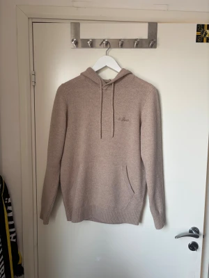 Beige stickad hoodie från MC2 Saint Barth - Säljer en beige stickad hoodie från MC2 Saint Barth med broderad logga på bröstet. Tröjan har huva med snörning, känguruficka och ribbade muddar. Perfekt för dig som gillar en clean och chill stil.