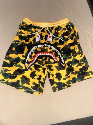 Gula camouflage shorts med hajmotiv - Säljer ett par gula shorts med grönt och svart camouflage-mönster och ett stort hajansikte framtill. Shortsen har resårmidja med vit snörning och är tillverkade i ett syntetmaterial som passar perfekt till sommaren.