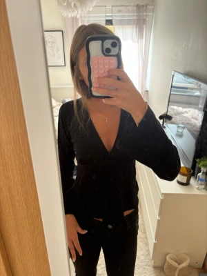 Svart omlottopp med knyt i midjan - Snygg svart omlottopp med v-ringning och knyt i midjan. Toppen har långa ärmar och en figurnära passform som framhäver midjan. Perfekt att styla med jeans eller kjol för en trendig look.