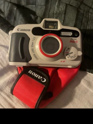 Canon Prima AS-1 kompaktkamera - Canon Prima AS-1 är en kompakt analog kamera med 32mm f/3.5-objektiv. Kameran är grå med svarta och röda detaljer och har en robust plastkonstruktion. Den levereras med ett rött Canon-fodral och har inbyggd blixt samt AiAF-autofokus. Kameran är Otestad och har en liten defekt se bild 4