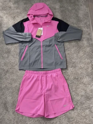 Nike miller set  - Snyggt träningsset från Nike med en rosa och grå jacka med huva och dragkedja samt matchande rosa shorts. Jackan har svarta detaljer på axlarna och praktiska fickor med dragkedja. Perfekt för löpning eller gym. Pris inte hugget i sten. Storlek M passar S och L