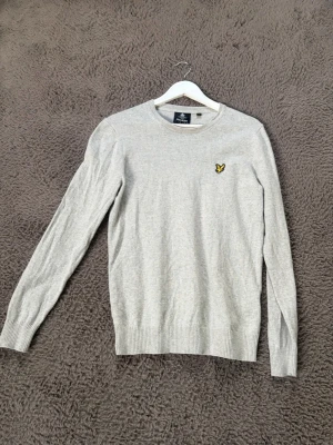Ljusgrå stickad tröja Lyle & Scott - Snygg ljusgrå stickad tröja från Lyle & Scott med rund halsringning och den klassiska gula loggan broderad på bröstet. Tröjan har långa ärmar och ribbade muddar vid ärmslut och nederkant. Perfekt till lager på lager och enkel att matcha.