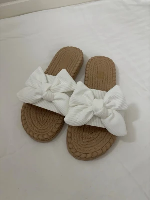 Beige sandaler med stora vita rosetter - Oanvända! Snygga sandaler med flätad beige sula och breda vita band dekorerade med stora rosetter. Perfekta för sommaren och ger en söt touch till din outfit. Tillverkade i textil och syntetmaterial med öppen tå och slip-in modell.