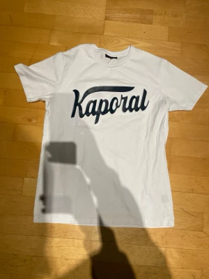 Vit t-shirt från Kaporal - Säljer en vit t-shirt från Kaporal med stor svart logga framtill. Klassisk rund hals och korta ärmar. Tillverkad i mjuk bomull som känns skön mot huden. Perfekt till jeans eller shorts för en clean och enkel stil.
