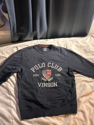 Mörkblå sweatshirt Polo Club Vinson - Mörkblå sweatshirt från Vinson Polo Club med rund halsringning. Framtill finns broderad text 'POLO CLUB' och ett amerikanskt sköldmärke i rött, vitt och blått. Tröjan har ribbade muddar vid ärmslut och nederkant. Perfekt för en sportig och avslappnad stil.