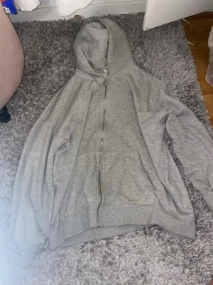 Grå zip hoodie med huva - En enkel och stilren grå hoodie med dragkedja framtill och stor huva. Hoodien har ribbade muddar vid ärmslut och nederkant samt två fickor framtill. Perfekt för en avslappnad och bekväm look. Materialet är mjukt och känns skönt mot huden.