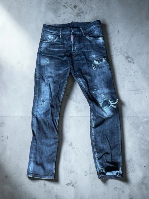 Svarta slitna jeans från Dsquared2 - Säljer ett par svarta jeans från Dsquared2 med slitningar och trasiga detaljer på benen. Jeansen har fem fickor, låg midja och en smal passform. Materialet är klassisk denim med en tvättad look och tydliga slitningar för en cool streetstil.