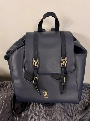 US Polo Assn. Mörkblå ryggsäck med gulddetaljer - Snygg mörkblå ryggsäck från US Polo Assn.  i slätt konstläder med guldfärgade spännen och logga framtill. Väskan har två justerbara axelremmar och praktiskt lock med dubbla remmar. Perfekt för dig som vill ha en stilren och trendig look.