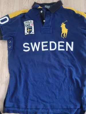 Marinblå pikétopp Sweden Polo - Pikétröja från Polo by Ralph Lauren . Tröjan har en stor gul broderad Ralh lauren på bröstet och texten "SWEDEN" i vitt. På vänster/höger arm finns siffran "10" i vitt . Baksidan av tröjan har texten "SWE" och siffran "10" i vitt. Har en slits på nedre delen. Men inga funna defekter . Klassisk krage och knappar framtill. Tillverkad i bomull. Tveka inte att höra av dig vid frågor!