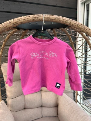 Tröja för barn - Söt rosa långärmad topp med tryck av tre får och texten 'Three in a row...' framtill. Rund halsringning med tryckknappar på axeln för enkel påklädning. Liten svart etikett nertill. Perfekt för dig som gillar gulliga motiv!