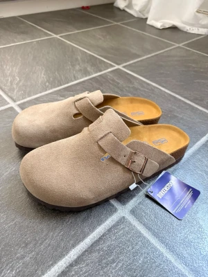 Birkenstock Boston - Säljer ett par snygga beige Birkenstock bostons i mjuk mocka. Modellen har en justerbar rem med metallspänne och klassisk korksula för extra komfort. Perfekta för dig som gillar stilrena och bekväma skor. Dem är helt nya. Skick 10/10