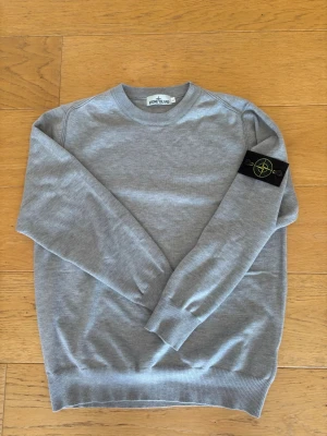 Grå sweatshirt från Stone Island - Snygg grå stickad sweatshirt från Stone Island med klassisk rund hals och ribbade muddar. Tröjan har den ikoniska svarta patchen med gul och grön kompasslogga på vänster ärm. Perfekt för en clean och stilren look. Är st XL men sitter som L!!