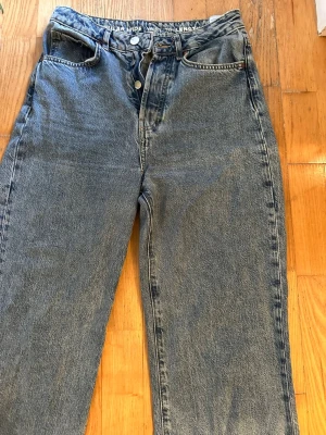 Blåa raka jeans med hög midja - Säljer ett par klassiska blå jeans med rak passform och hög midja. Jeansen har knappgylf och fem fickor. Materialet är kraftig denim i bomull och färgen är en tvättad blå nyans. Perfekta för en avslappnad och trendig look.